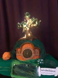 A pumpkin covered in felt and felting wool to depict Bag End. The stem has been turned into a tree that is lit up with lights. There is a round door colored on the front and a sign that reads 'No Admittance Except on Party Business' in the front next to a mossy fence. The garden has small green shrubs and a bush covered in white flowers. There is also a small needle felted jack o lantern in the garden. There is a purple curtain in the background.