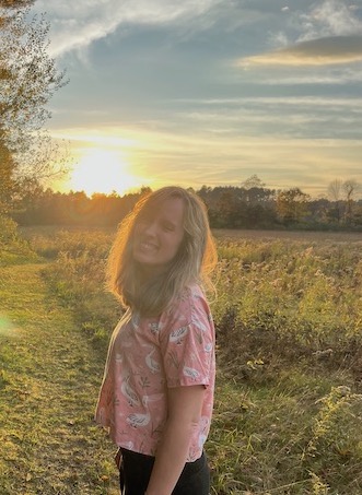 Rebecca Doris, standing on a mowed path on the border of meadows, with a line of trees in the background and a golden sunset shining into the camera. Rebecca is smiling and laughing, eyes closed.